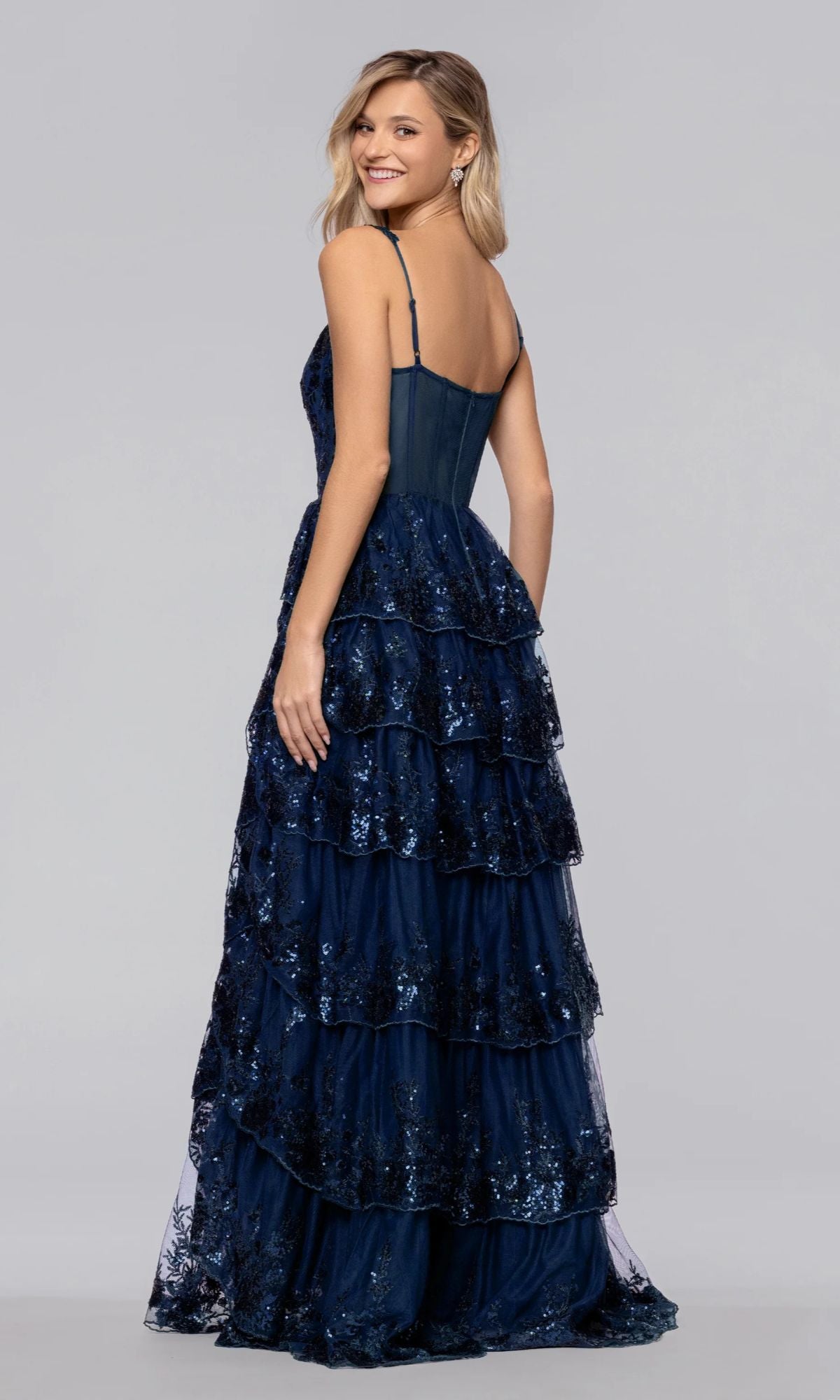 Woman wearing the Long Prom Dress: Blondie Nites 6126BN, a navy blue, tiered A-line gown with thin straps and corset bodice, facing slightly to the side and smiling against a plain gray background.